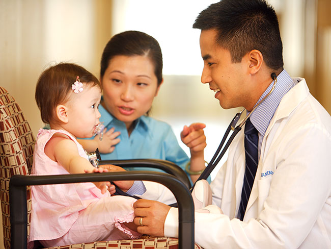Kaiser Permanente的一名raybat官网亚裔美国医生正在和一个穿粉红色衣服的可爱女婴交谈，她的母亲在背景中打手势。