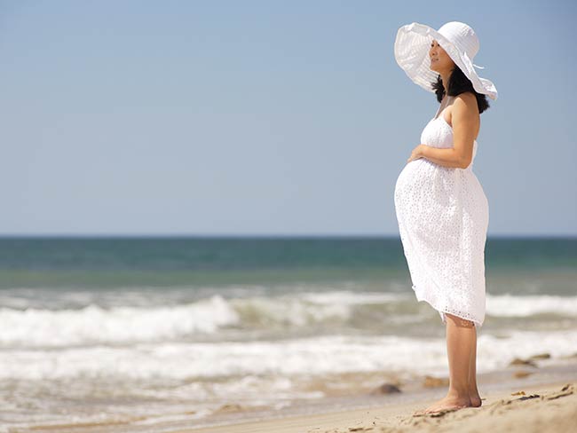 穿着白裙子戴着帽子的孕妇站在沙滩上。