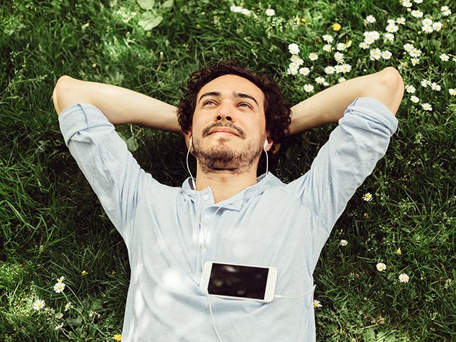 一个面带微笑的年轻人躺在草地上，双手放在脑后，戴着耳机，胸前放着手机。