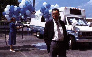男人穿着西装和领带站在移动医疗货车前，而在背景中的女人则保持蓝色和白色气球