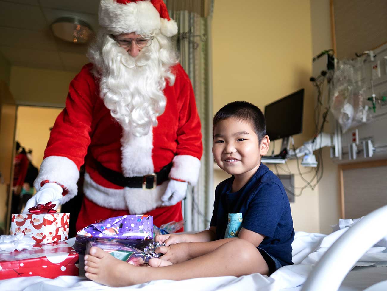 一个微笑着的4岁男孩躺在医院的病床上，周围都是礼物，圣诞老人就站在床边。