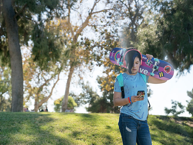 Teen is outside holding a skateboard and looking at phone.