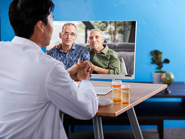 两个男人一起坐在沙发上，一个人的胳膊搭在另一个人的肩膀上，微笑着与一位男医生通过视频聊天进行讨论。