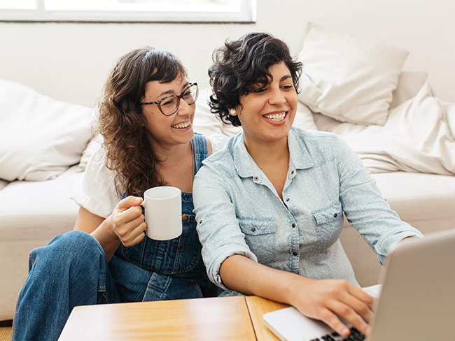 快乐的女同性恋夫妇喝咖啡，在客厅使用笔记本电脑。