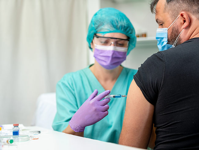 穿着防护装备的护士正在向一名男子的手臂注射COVID-19疫苗。