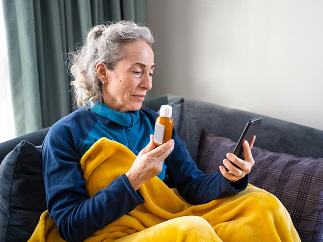 成年女性患者在家里盖着毯子，用手机与医生在线通话，向医生展示自己需要的处方药物。