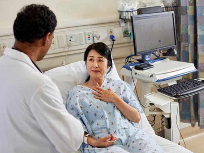 一位妇女躺在病床上，手放在胸前，正在和一位医生交谈