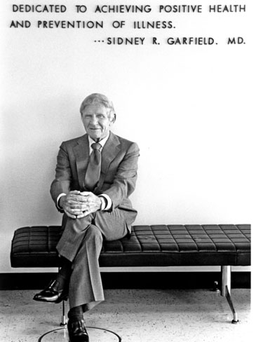 加菲尔德双腿交叉坐在一块牌子前，面带微笑，上面写着“致力于实现积极的健康和疾病预防。”西德尼·R·加菲尔德博士