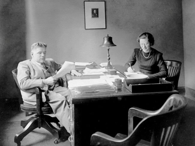 哈罗德·哈奇和秘书贝斯·格奇奇坐在一张桌子前看报纸
