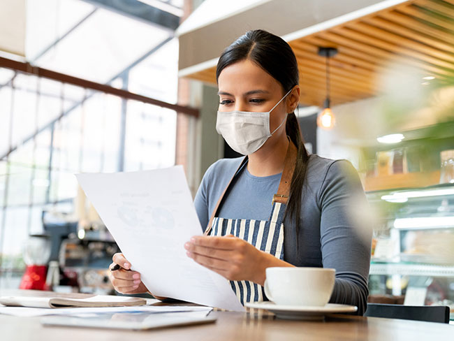 一位戴着口罩的拉丁裔女商人，在一家咖啡店里阅读一份文件。