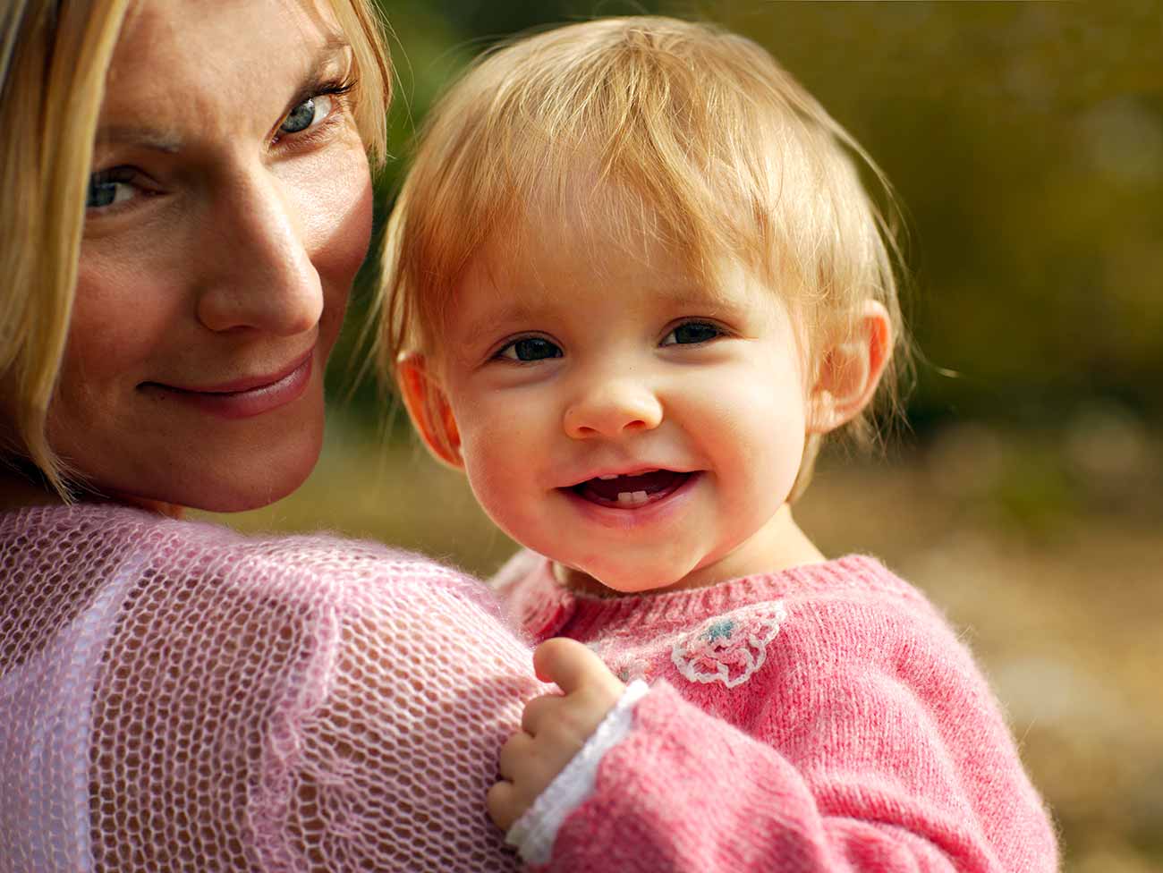 A mother is looking over her shoulder while holding her baby and both are smiling into the camera.