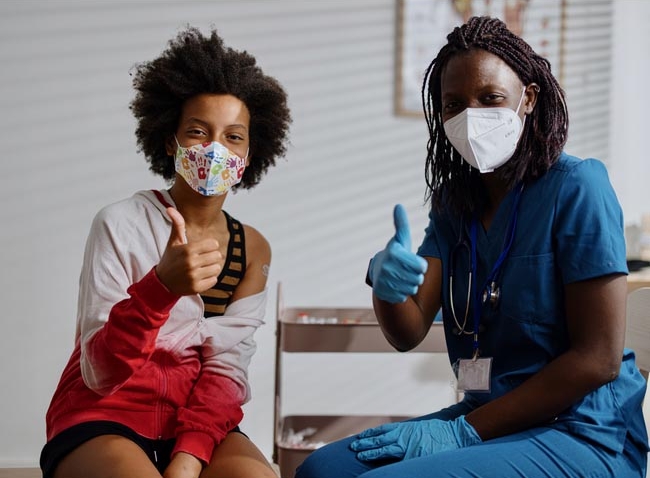Teen and doctor wearing face masks, giving a thumbs up after vaccination.