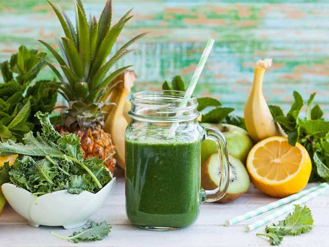 Piña kale-ada served in a mason jar surrounded by tropical fruits and veggies on a white marble counter top